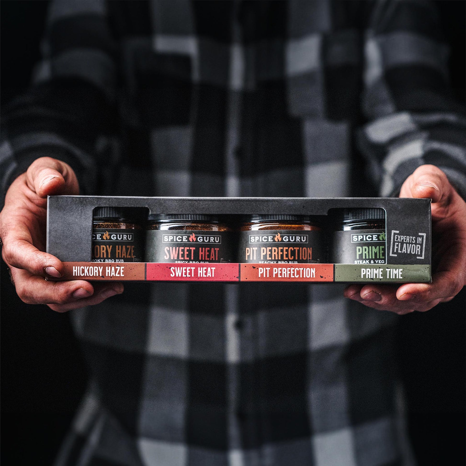 Extended hands holding a box with containers of Spice Guru Pit Perfection, Sweet Heat, Prime Time, and Hickory Haze BBQ Rubs.