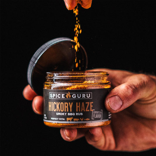 A hand holding an open container of Spice Guru Hickory Haze: Smoky BBQ Rub, sprinkling spices into it.