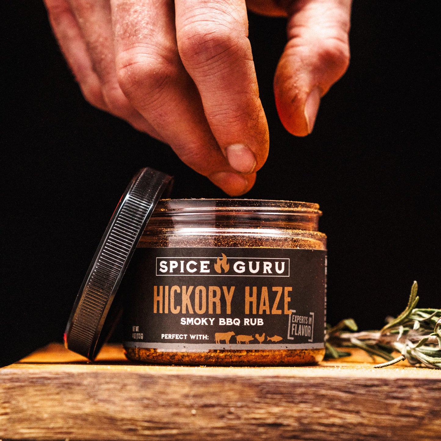 A hand reaching towards an open container of Spice Guru Hickory Haze: Smoky BBQ Rub, sitting on a wooden table with fresh herbs.