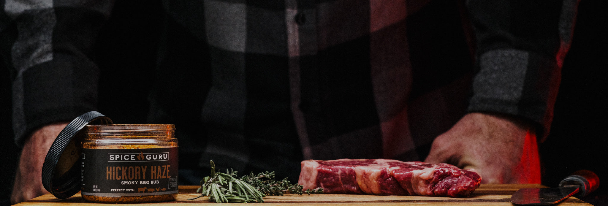 An open container of Spice Guru Hickory Haze, sitting on a butcher block with a steak, fresh herbs, and a chef's knife.