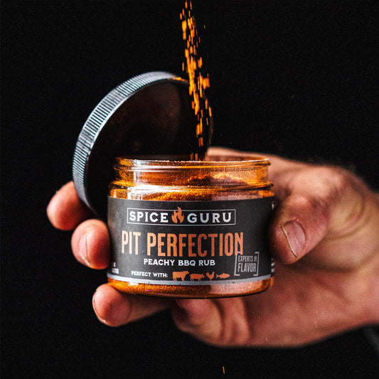 A hand holding an open container of Spice Guru Pit Perfection: Peachy BBQ Rub, sprinkling spices into it.