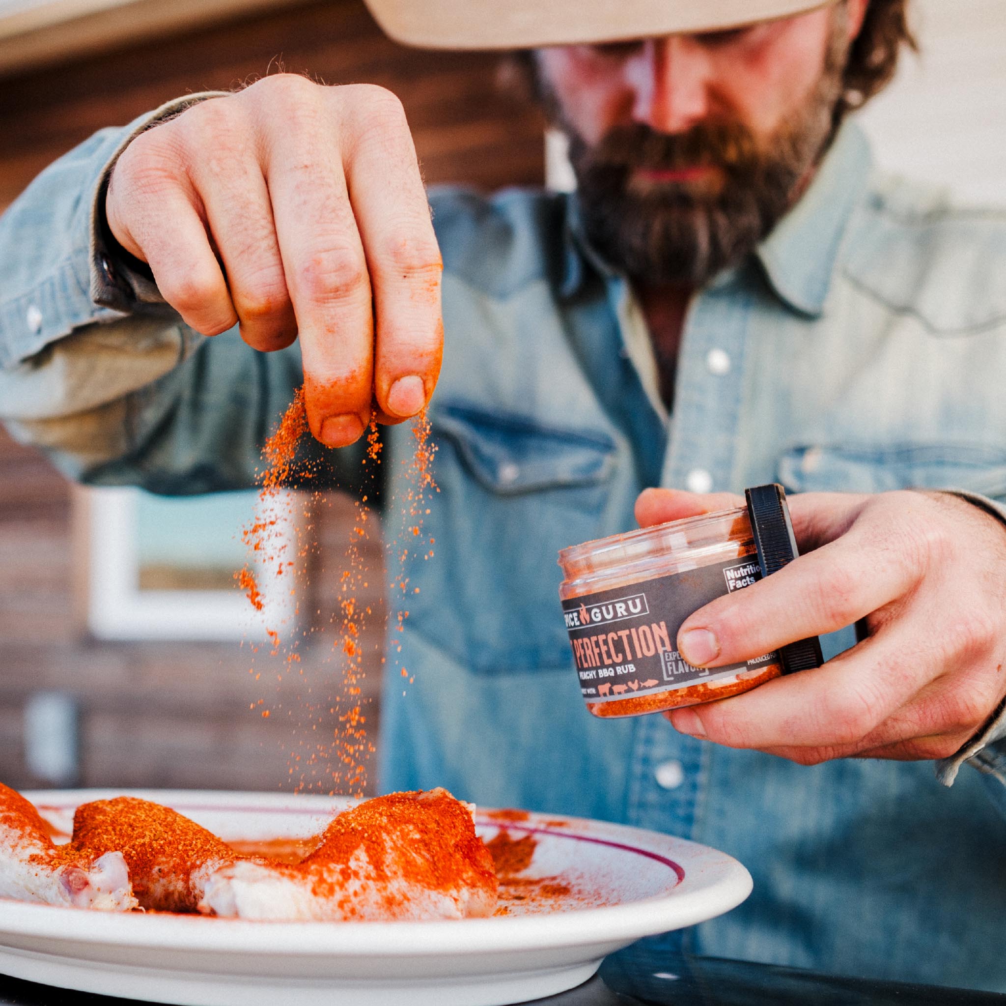 A bearded man in a blue shirt sprinkling Spice Guru Pit Perfection onto chicken drumsticks, holding the open container in the other hand.