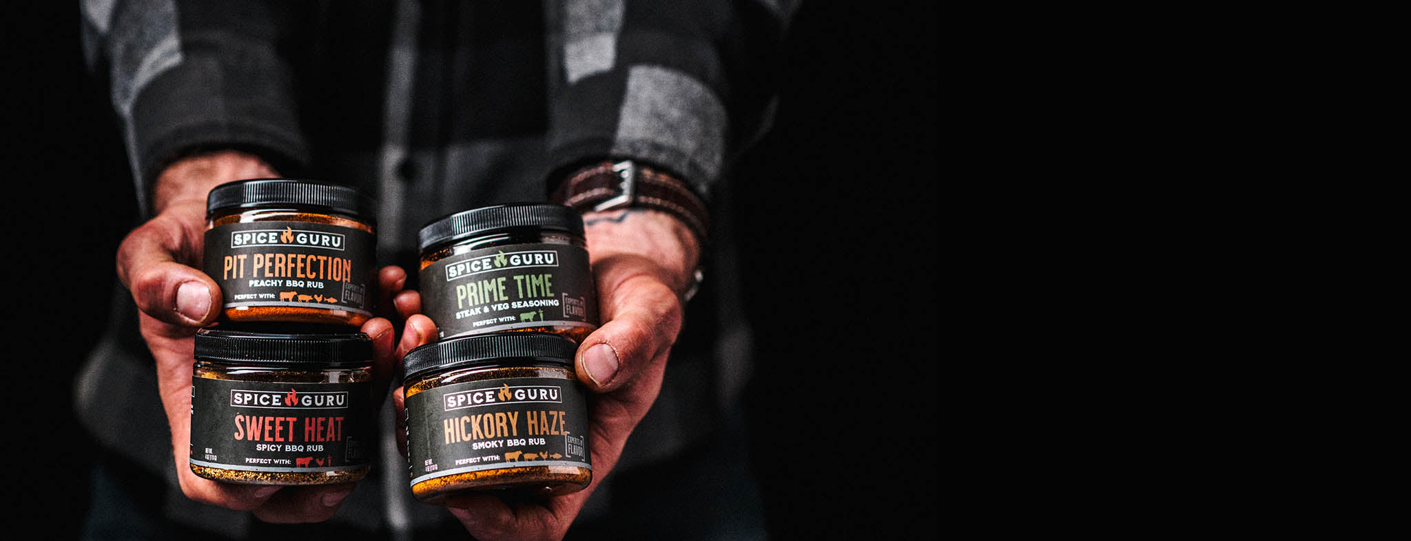 Extended hands holding containers of Spice Guru Pit Perfection, Sweet Heat, Prime Time, and Hickory Haze BBQ Rubs, two in each hand.