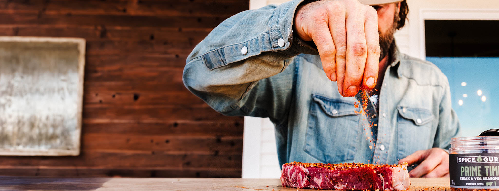 A bearded man in a blue shirt sprinkling Spice Guru Prime Time onto steak.