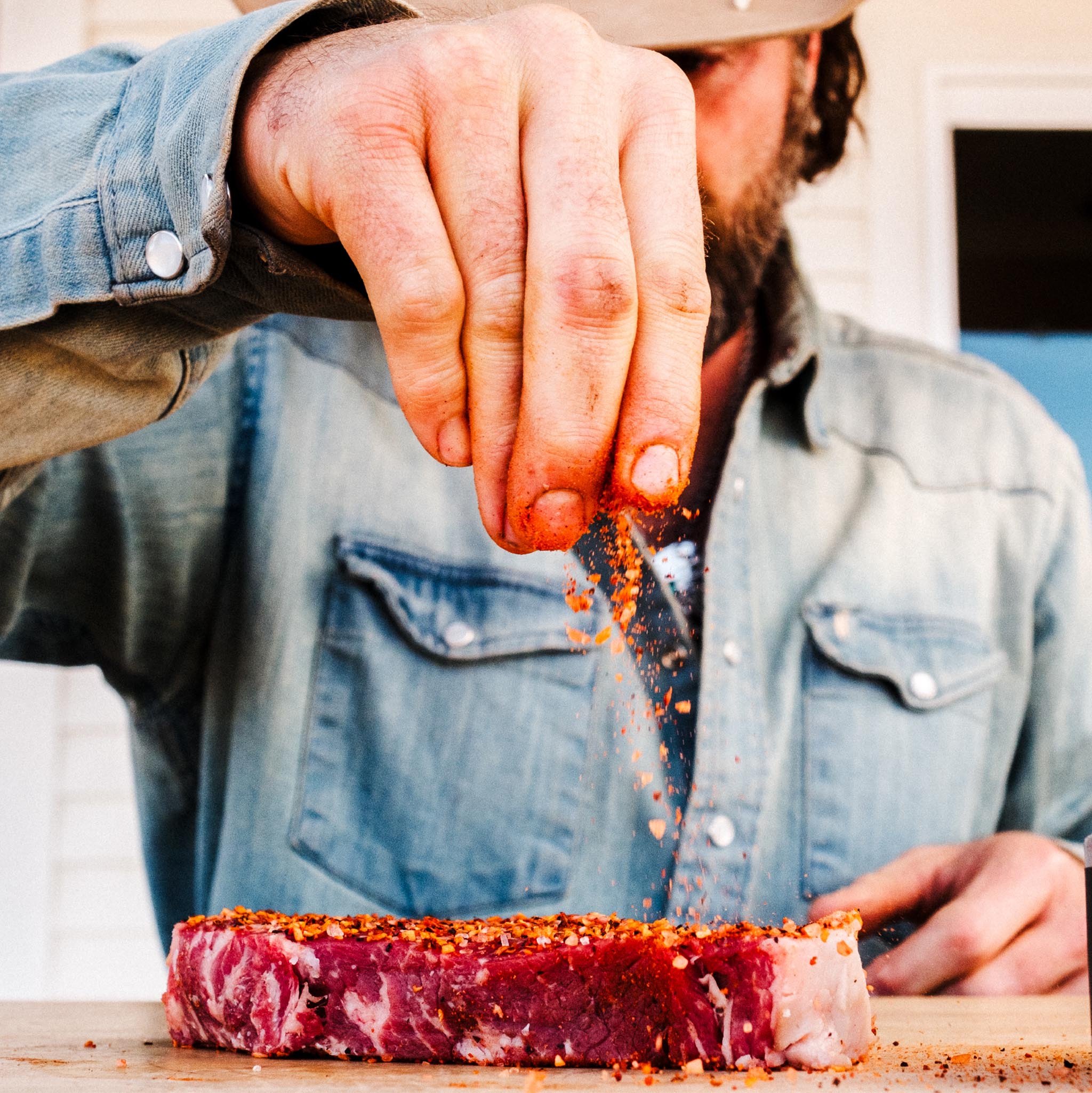 A bearded man in a blue shirt sprinkling Spice Guru Prime Time onto steak.