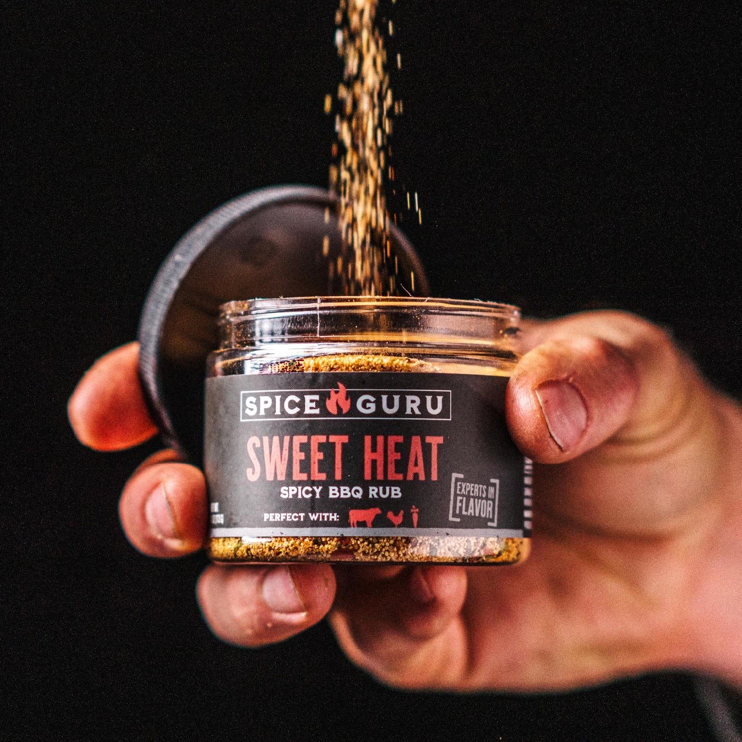 A hand holding an open container of Spice Guru Sweet Heat: Spicy BBQ Rub, sprinkling spices into it.