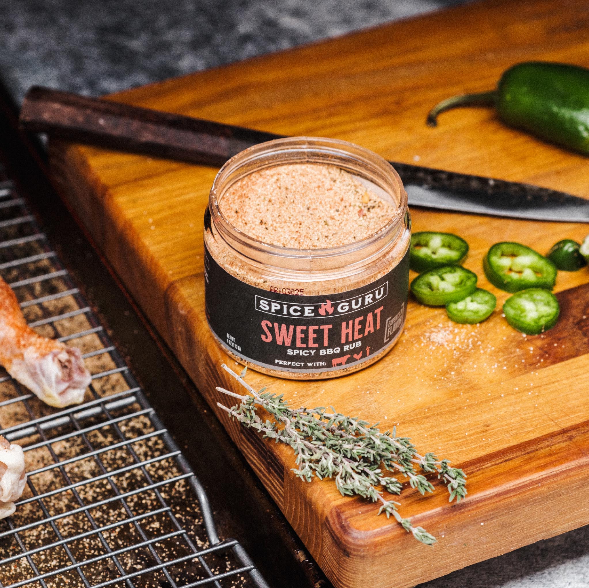 An open container of Spice Guru Sweet Heat, sitting on a wooden cutting board with fresh herbs, sliced peppers, and a chef's knife.