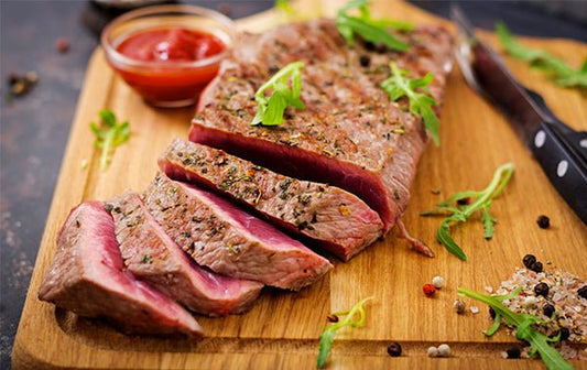 A carved steak sprinkled with fresh herbs, resting on a wooden cutting board with a chef's knife and a bowl of red sauce.